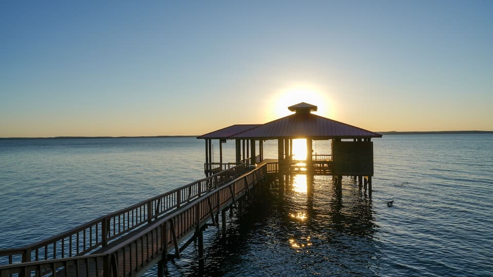 A pier extending into calm waters with the sun setting in the background.