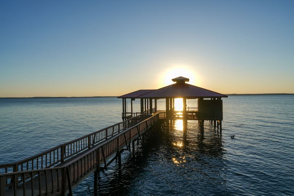 A pier extending into calm waters with the sun setting in the background.