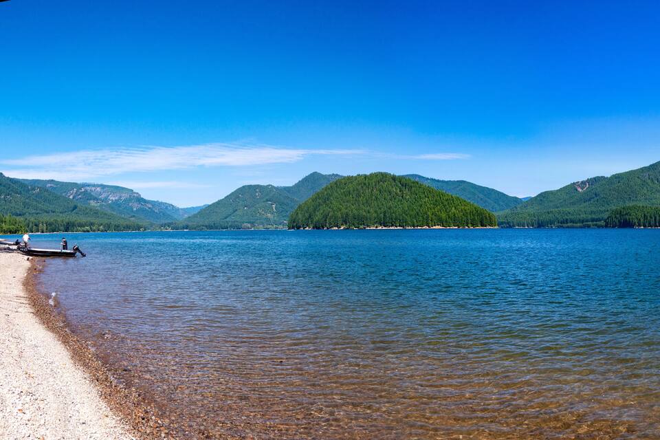 A calm lake with a boat on the shore, surrounded by forested mountains under a clear blue sky.