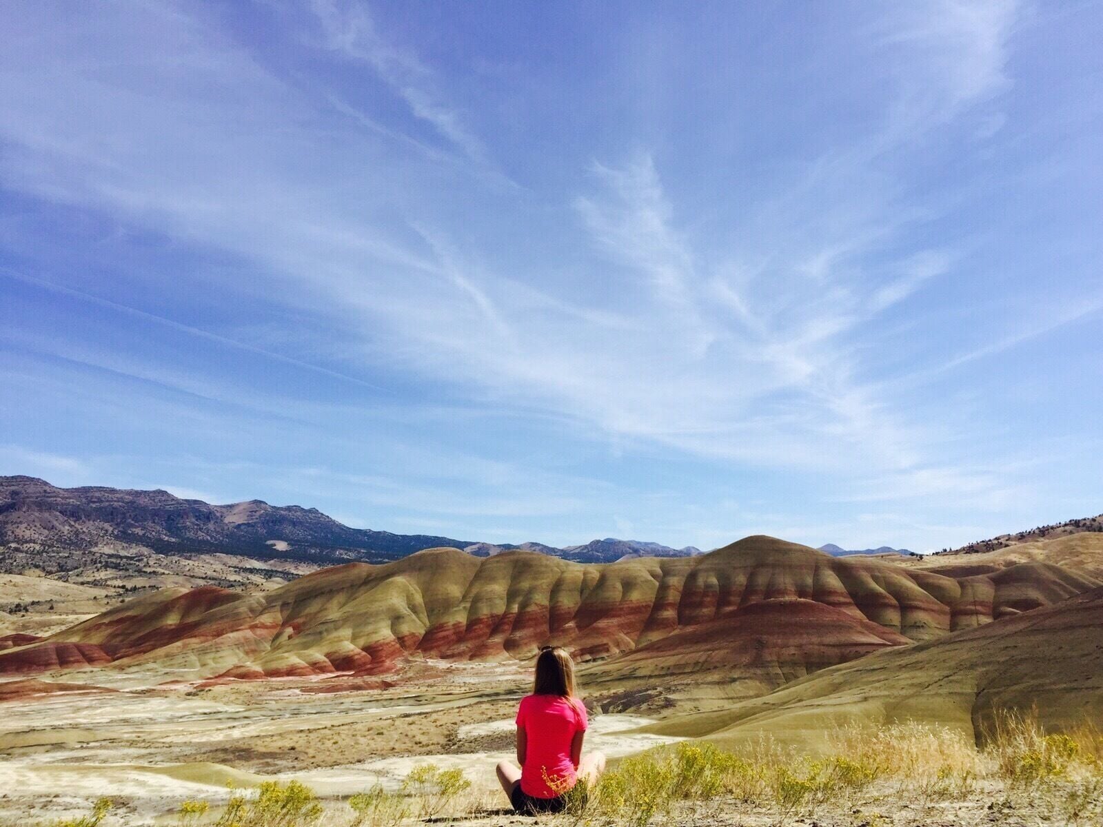 Uma pessoa sentada em uma colina, com vista para paisagens áridas coloridas e montanhas ao longe.