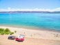 Une plage avec un camping-car rouge et une voiture garés sur le sable, un lac bleu limpide et des montagnes en arrière-plan.