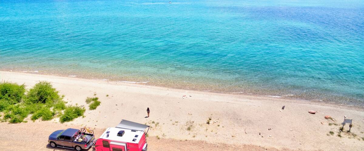 A beach with a red RV and a car parked on the sand, a clear blue lake, and mountains in the background.