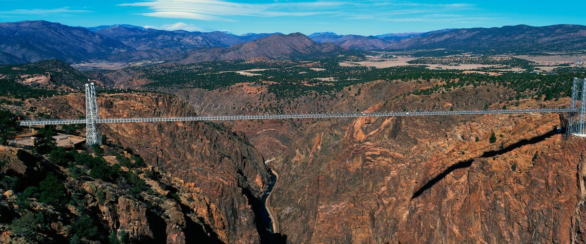 A suspension bridge spanning a deep canyon with rugged mountains in the background.