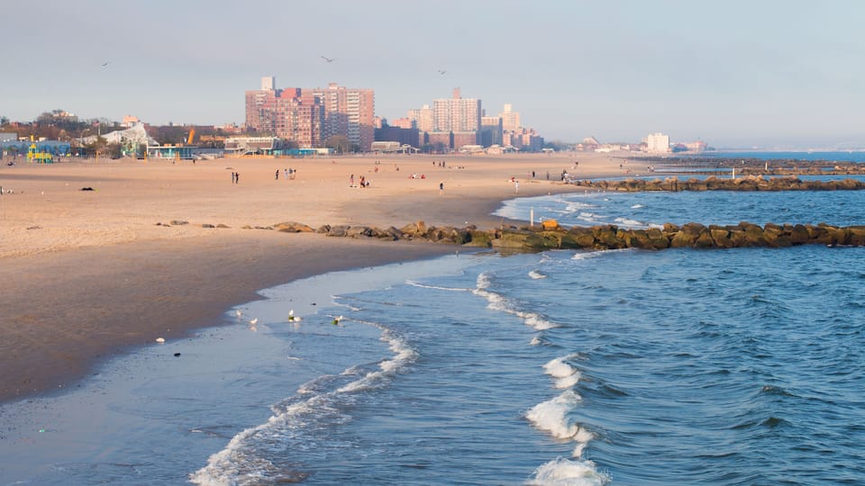 A beach with waves, a rocky breakwater, and buildings in the background.