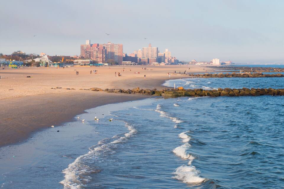 A beach with waves, a rocky breakwater, and buildings in the background.