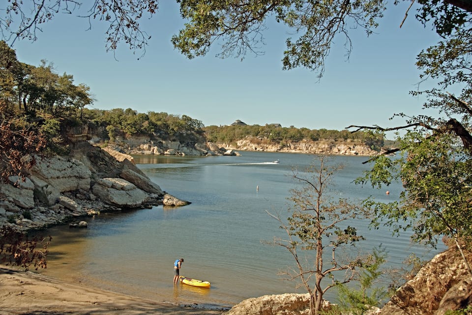 A calm river with rocky banks, a person in a blue shirt standing by a yellow kayak, and a boat in the distance.