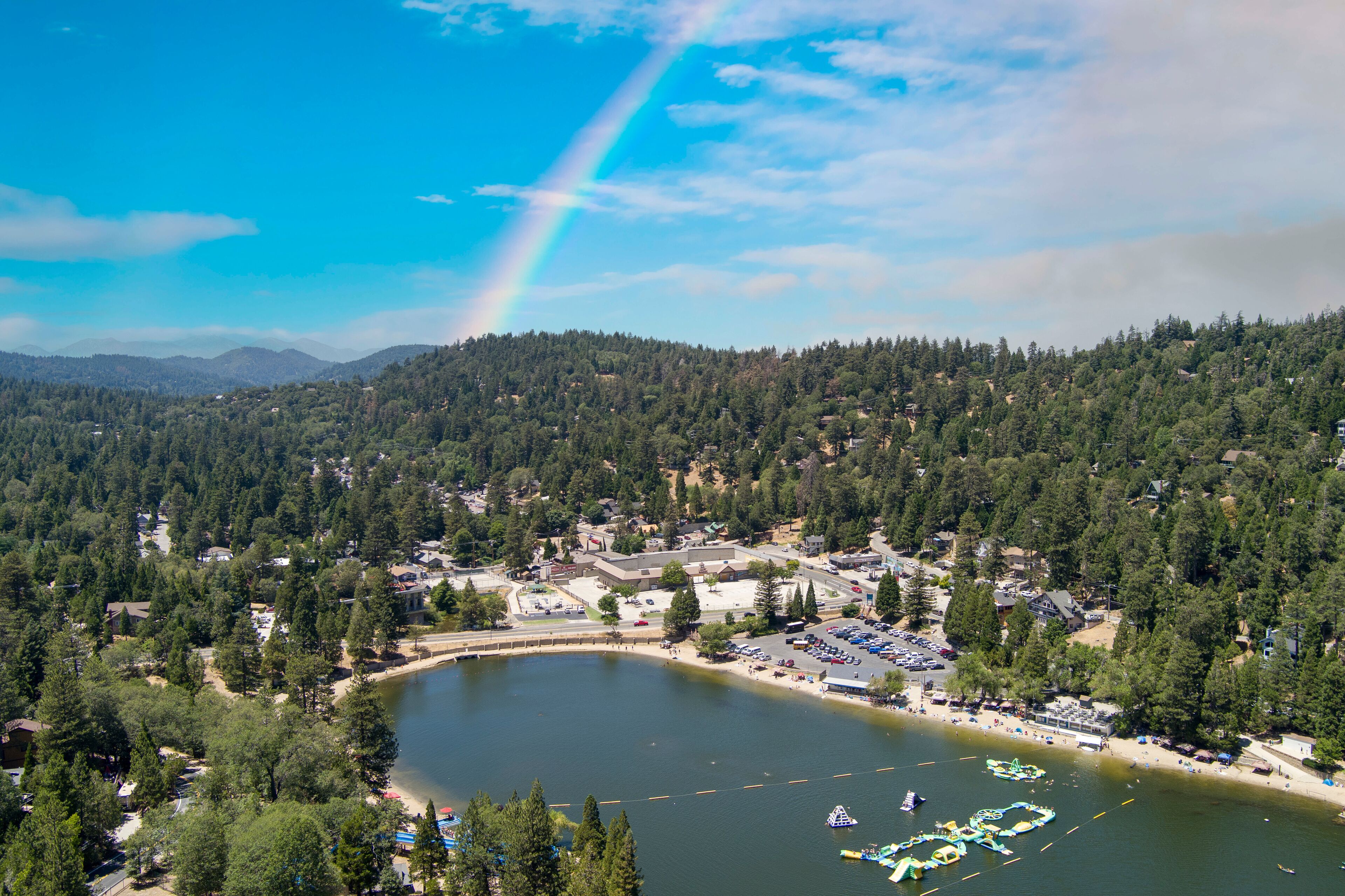 aerial shot of a gorgeous summer landscape with rippling lake water, green trees, grass and plants with homes on the hillside, blue sky, cloud sand a rainbow at Lake Gregory in Crestline California
