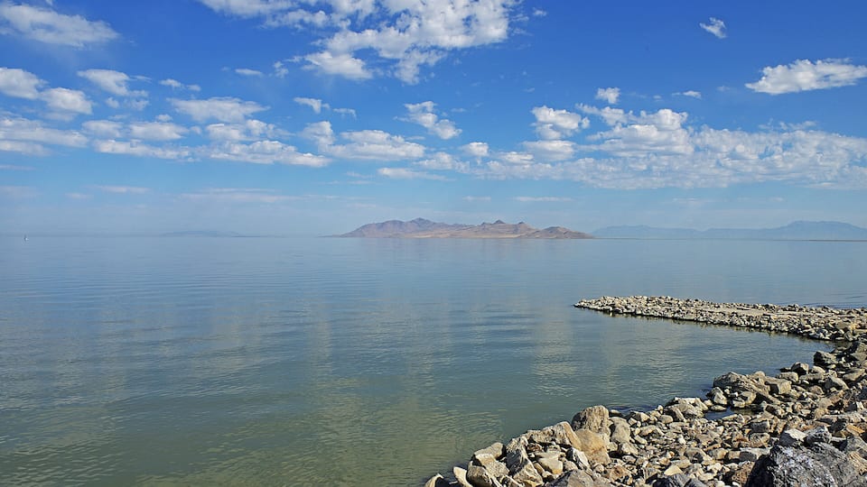 A calm lake with a rocky shoreline, clear blue sky, and distant mountains.