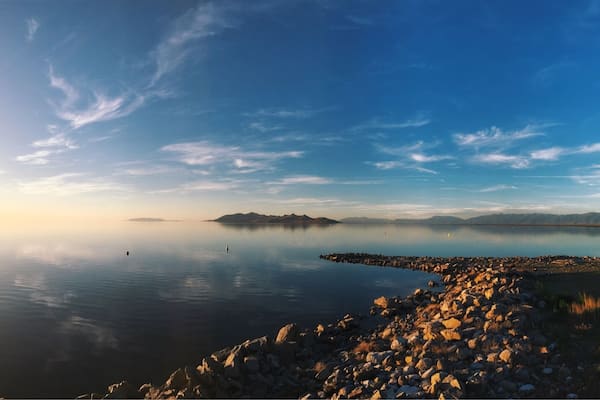 Un lac paisible, bordé d’une rive rocheuse, avec des montagnes lointaines qui se dessinent sous un ciel dégagé.