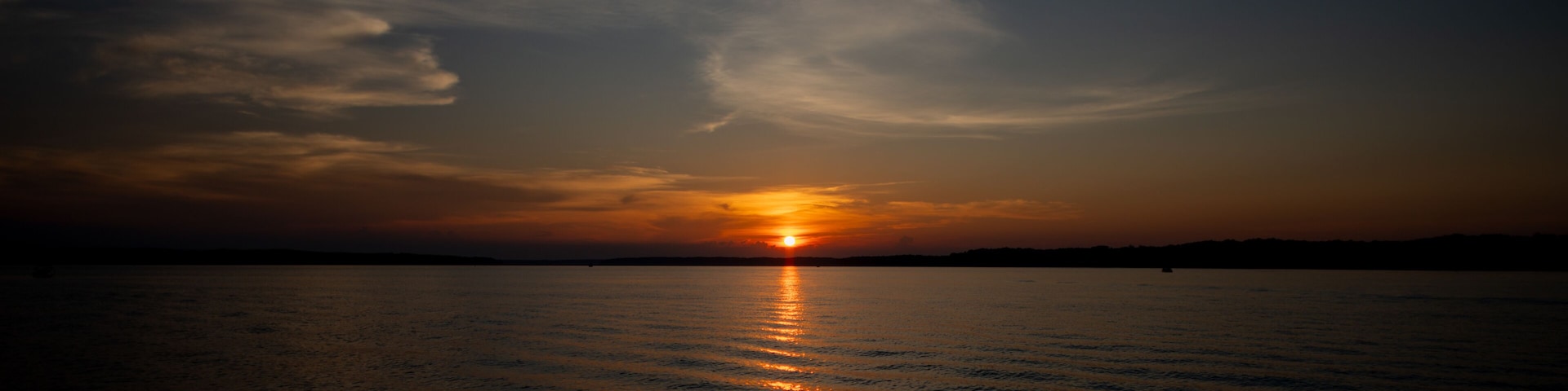 Un coucher de soleil sur une étendue d’eau calme, avec la silhouette d’une terre ferme à l’horizon.