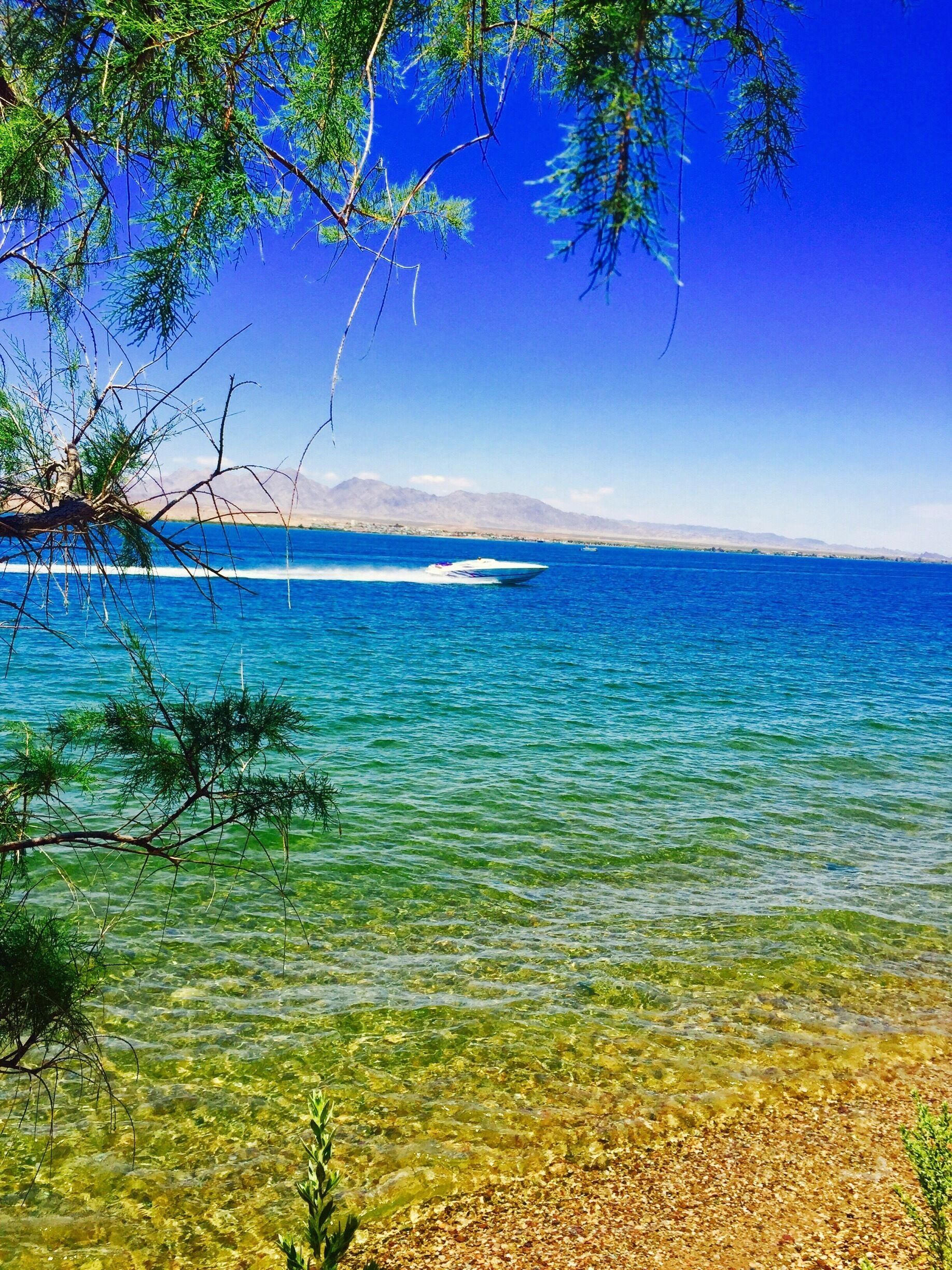 Une mer d’un bleu profond, avec un bateau, une plage de galets et des montagnes à l’horizon.