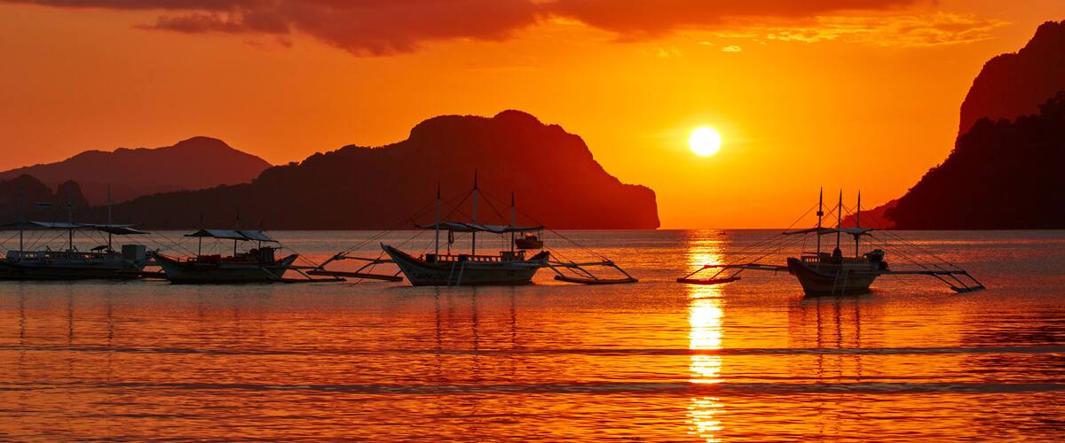 A sunset over a calm sea with boats and mountains in the background.
