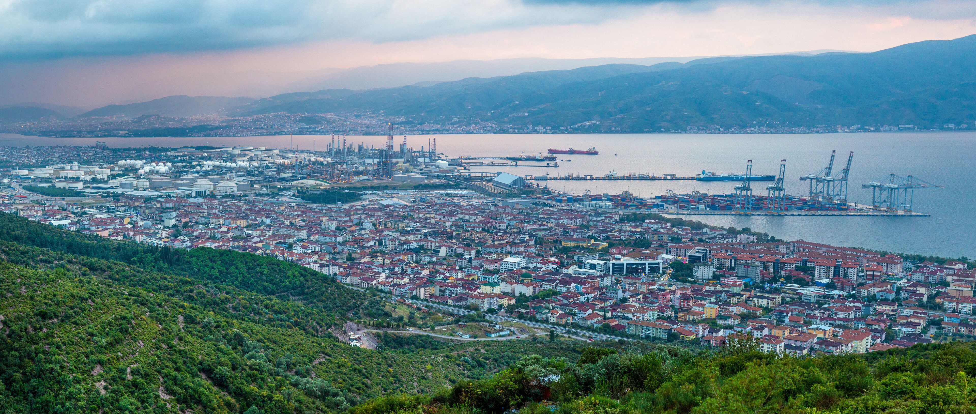 A coastal cityscape with dense urban buildings, a port with cranes, and a large ship in the water.