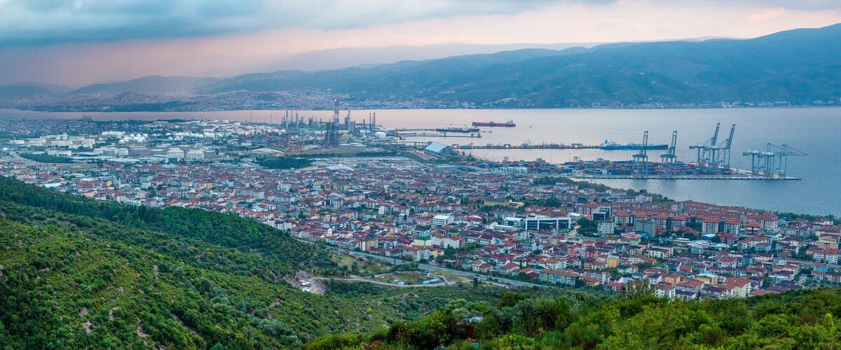 A coastal cityscape with dense urban buildings, a port with cranes, and a large ship in the water.