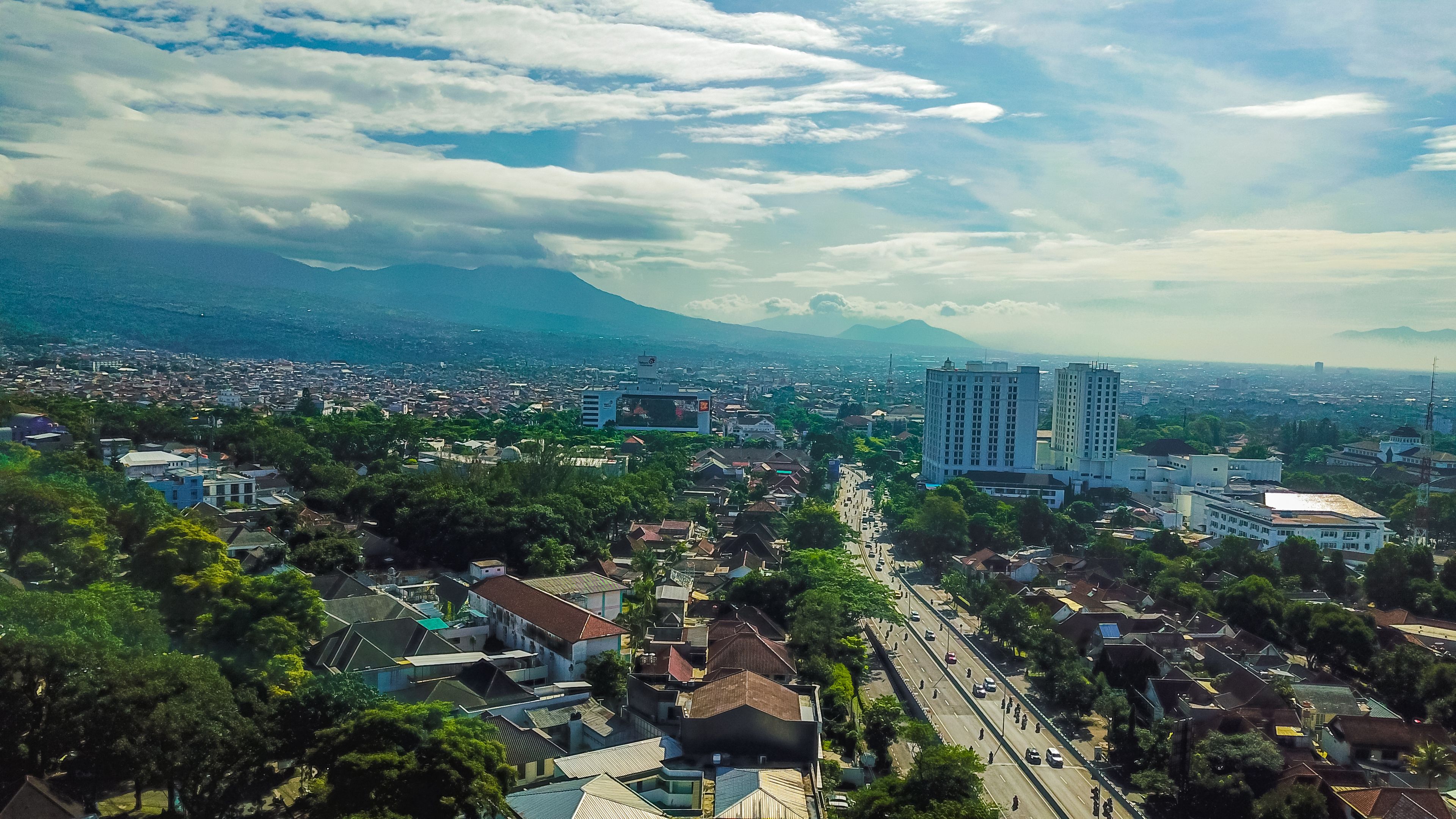 Un paysage urbain avec une montagne imposante en arrière-plan, une large route où circulent des véhicules et des quartiers résidentiels densément peuplés.