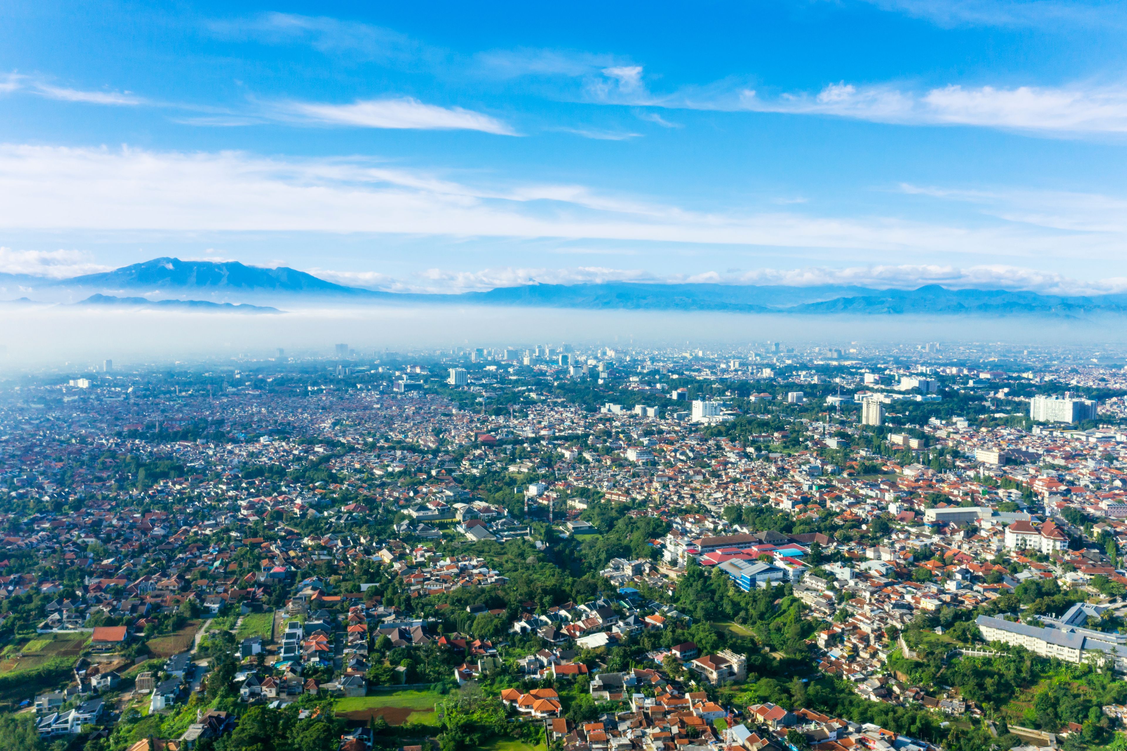 Une vue panoramique d’une ville avec des quartiers résidentiels densément peuplés, des espaces verts et des montagnes lointaines, le tout sous un ciel dégagé.