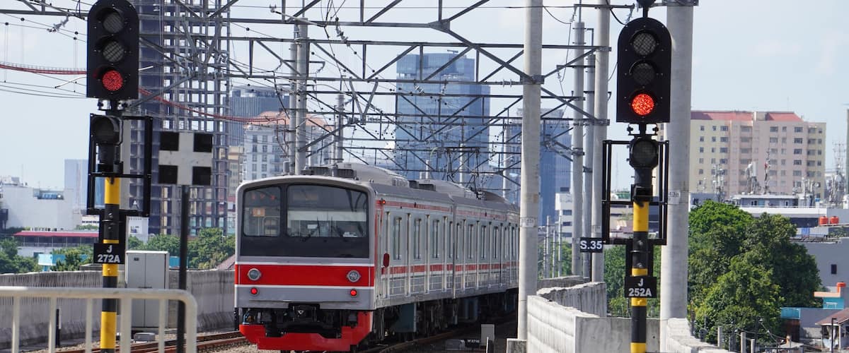Ein Zug passiert eine Bahngleisüberquerung, im Hintergrund sind Stromleitungen und Gebäude zu sehen.