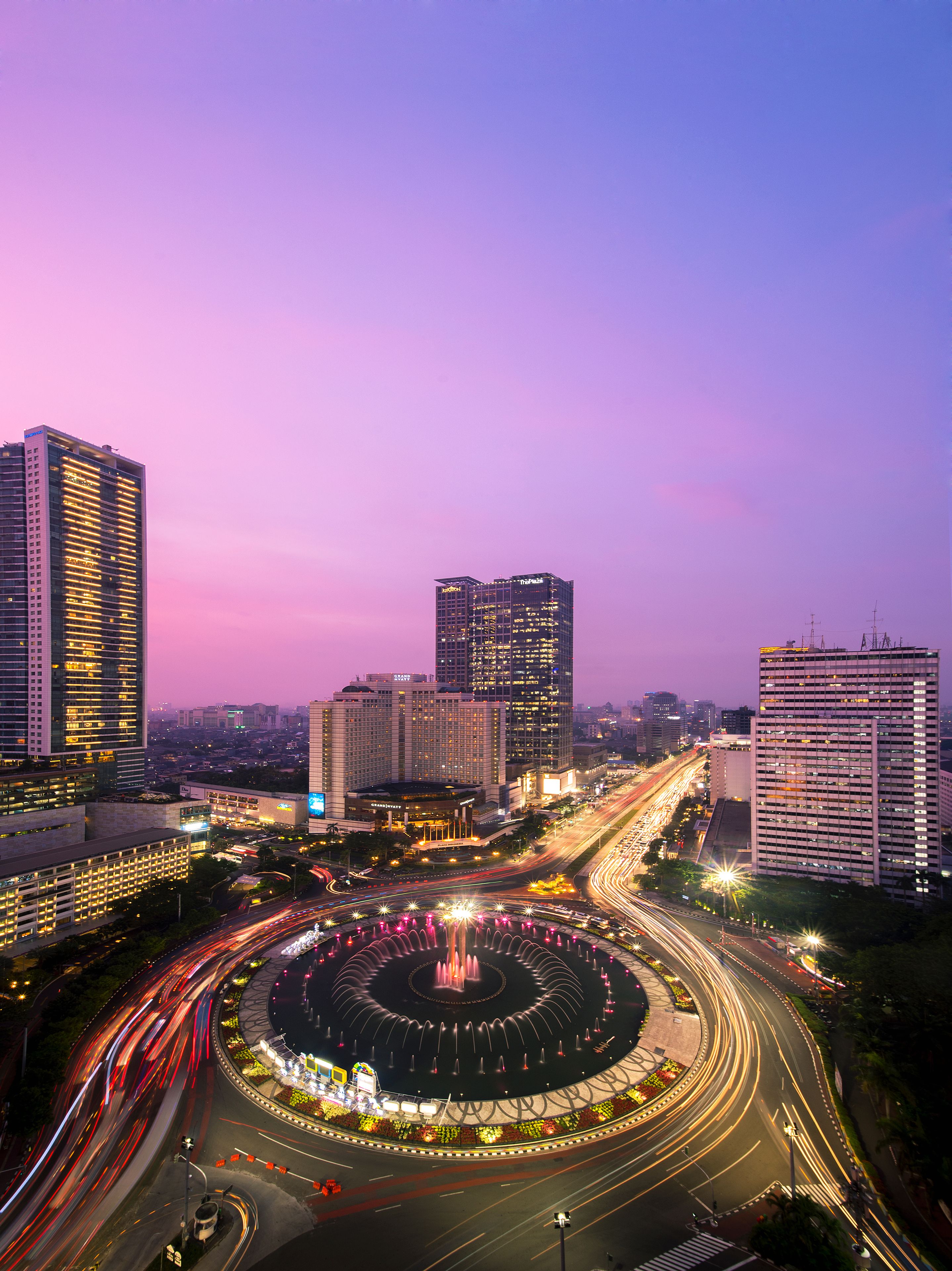 A cityscape at dusk with a circular water feature in the center, surrounded by high-rise buildings and busy roads.