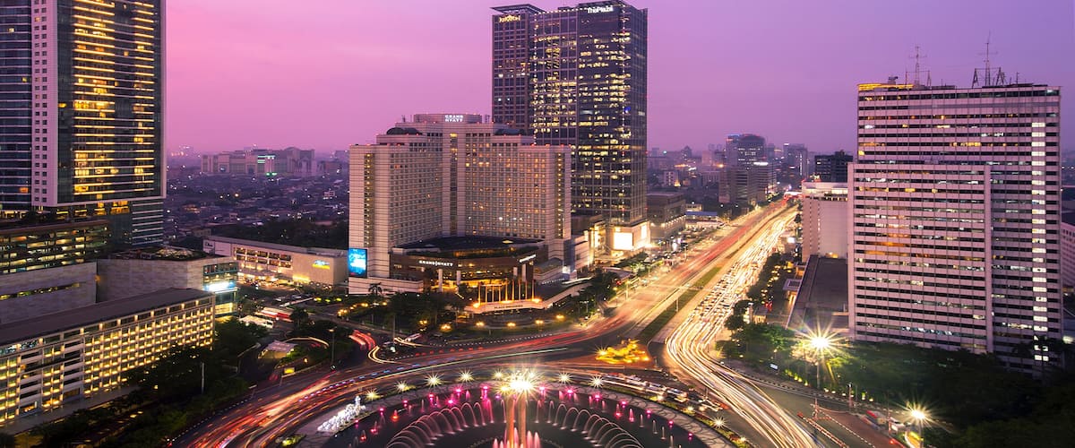Une vue sur la ville au crépuscule, avec une fontaine circulaire au centre, entourée de gratte-ciel et de rues animées.