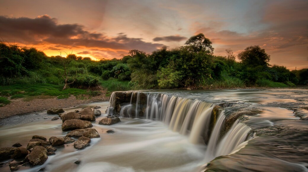 A waterfall cascading over rocks, surrounded by lush greenery and a sunset sky.
