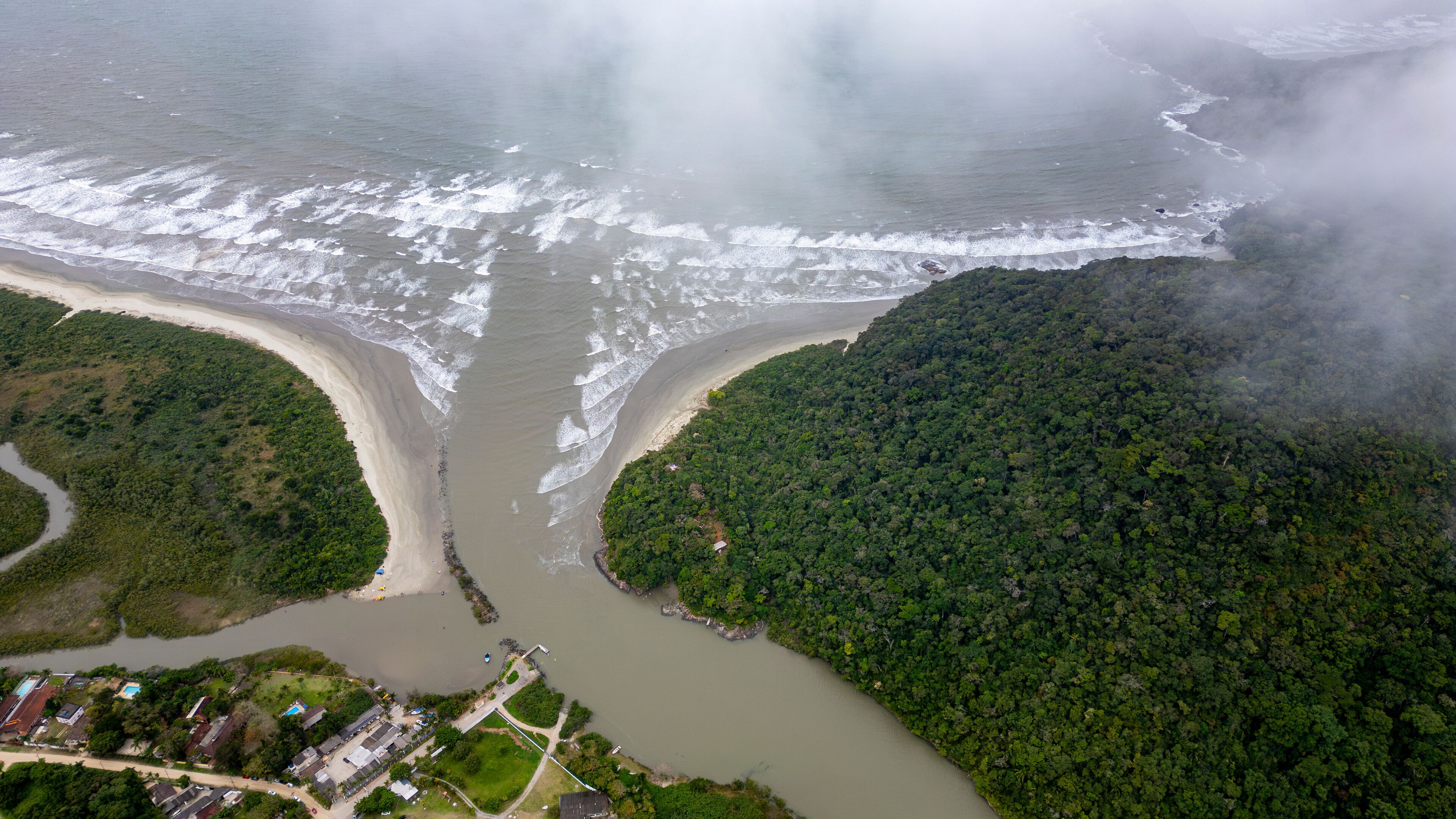 An aerial view of a coastal area with a river flowing through a forested peninsula, a beach, and a small settlement with buildings and roads.