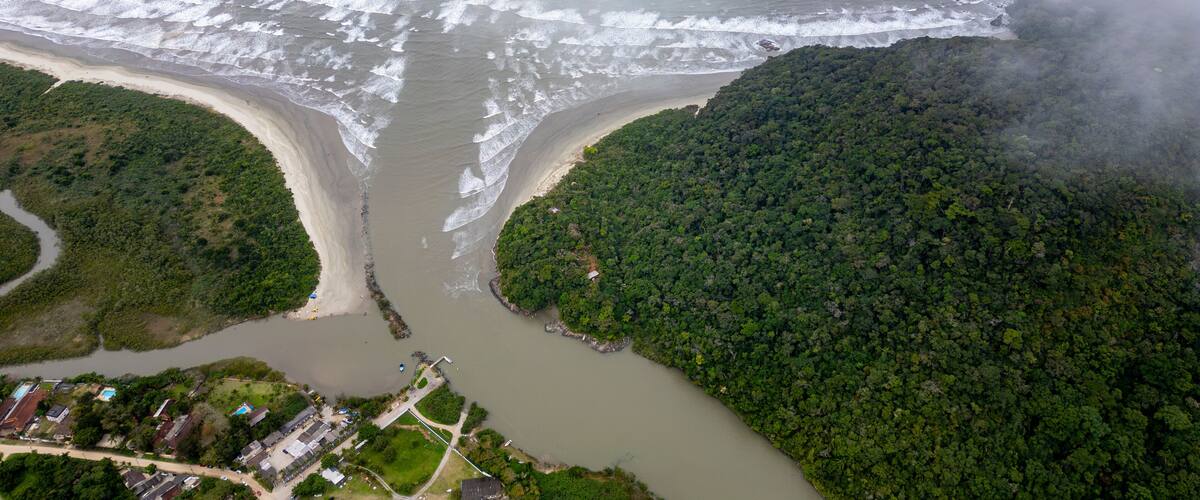An aerial view of a coastal area with a river flowing through a forested peninsula, a beach, and a small settlement with buildings and roads.