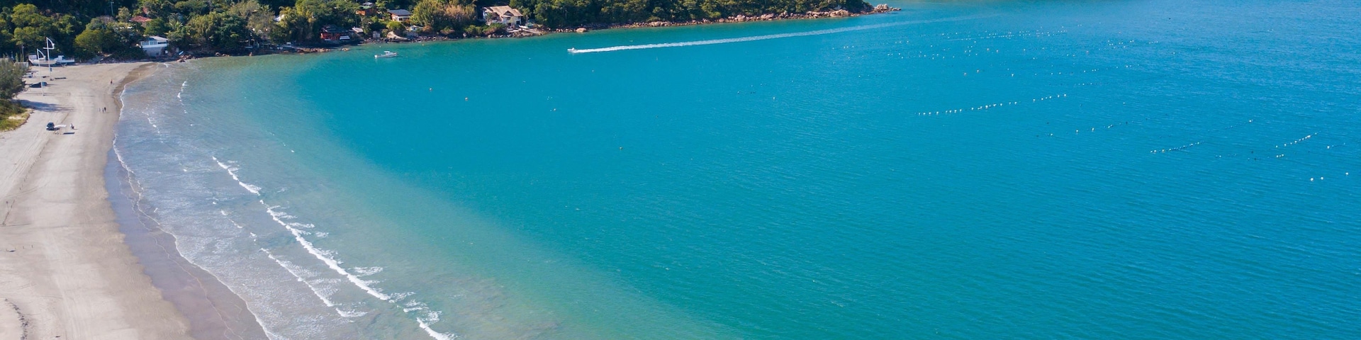 Paisagem costeira com uma praia arenosa, águas turquesa cristalinas e uma colina arborizada.