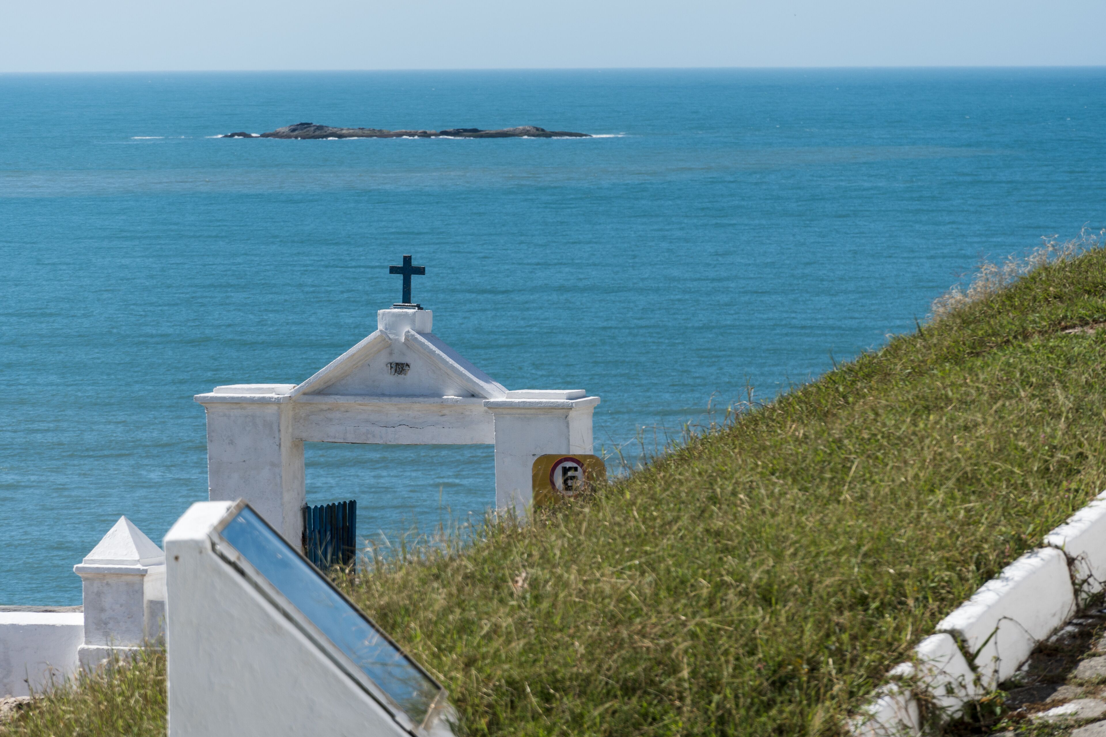 A white chapel with a cross on top, overlooking a calm sea with a rocky outcrop in the distance.