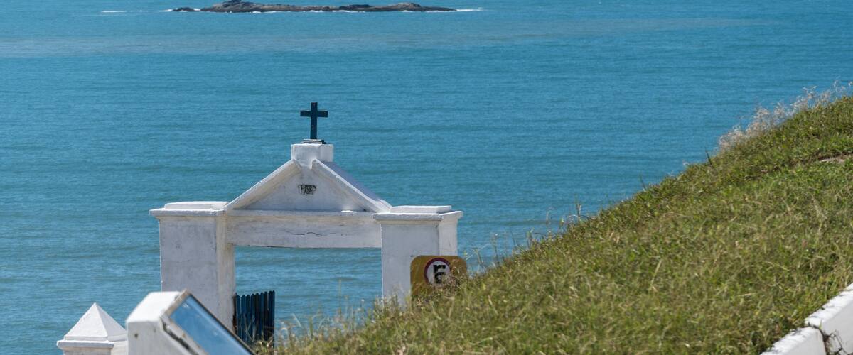 A white chapel with a cross on top, overlooking a calm sea with a rocky outcrop in the distance.