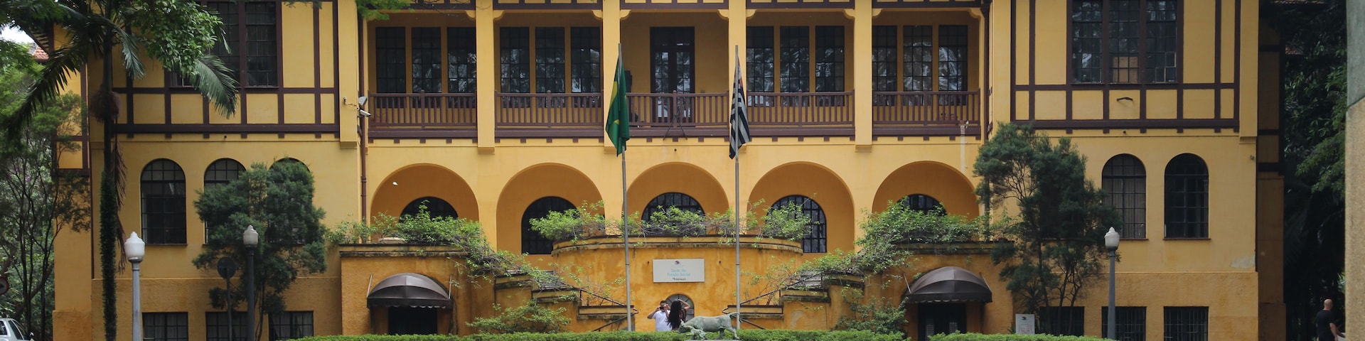 Um edifício amarelo com telhado de azulejos, rodeado por um gramado bem cuidado e árvores.