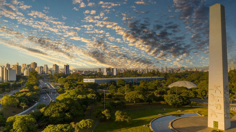 Paisagem urbana com um obelisco imponente, uma estrada sinuosa e um amplo parque verde com árvores.