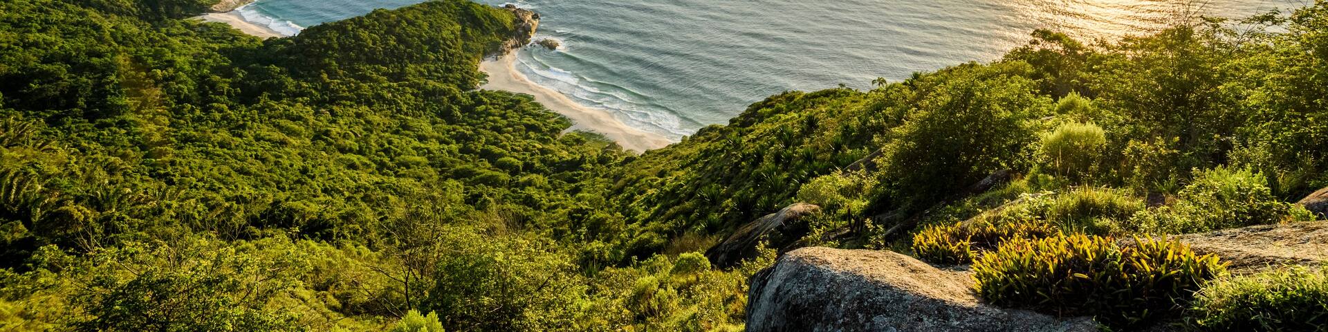 A coastal landscape with a clear sky, a sun setting or rising over the ocean, and a mountainous terrain with dense greenery.