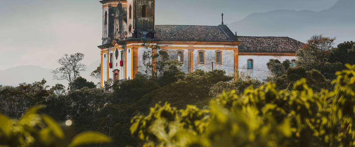 Une église historique dotée d’un clocher et d’un dôme, entourée d’une végétation luxuriante et avec des montagnes en toile de fond.