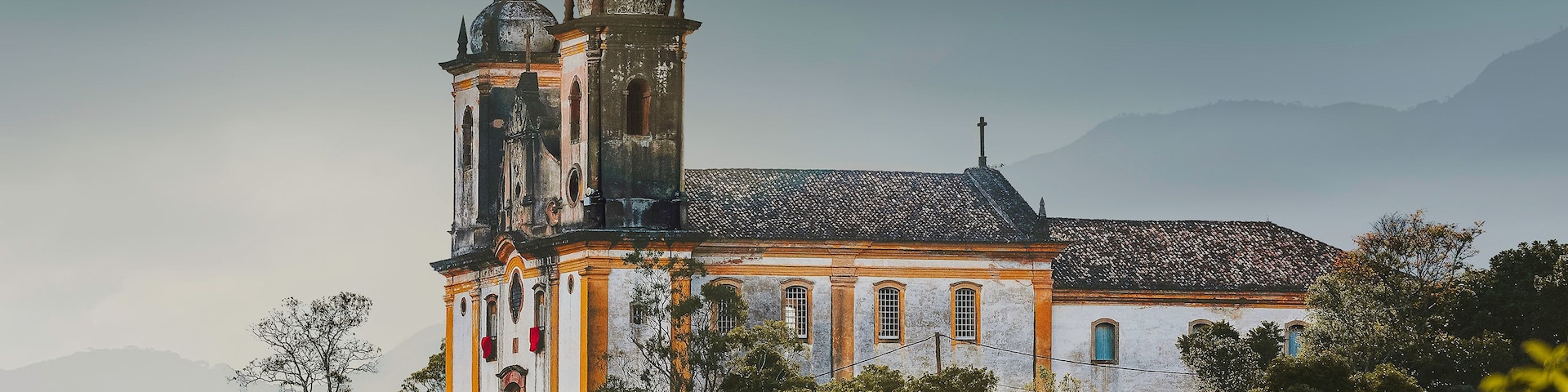 Une église historique dotée d’un clocher et d’un dôme, entourée d’une végétation luxuriante et avec des montagnes en toile de fond.