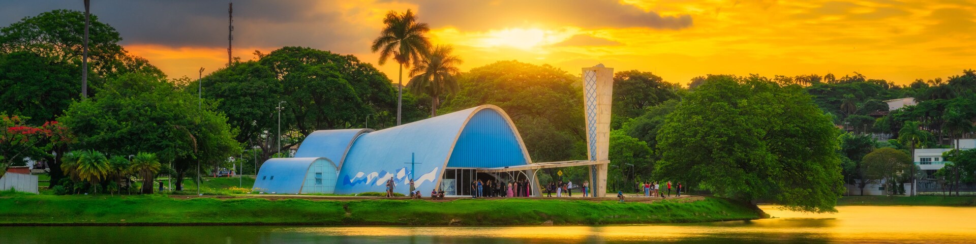 Estrutura azul com uma cruz, rodeada por árvores e um corpo de água refletindo o pôr do sol.