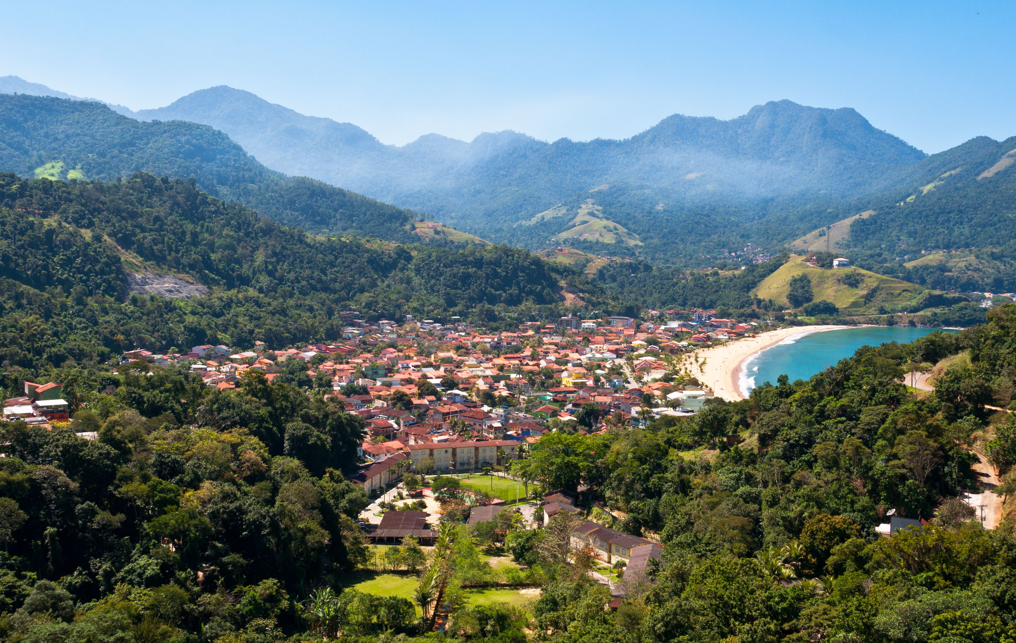 A coastal town with red-roofed buildings, a beach, and surrounding mountains.