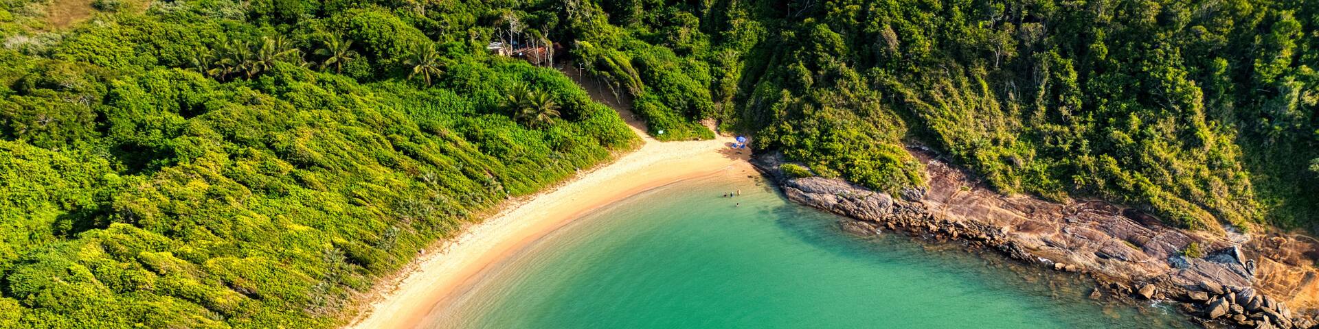 An aerial view of a coastal area with a sandy beach, clear turquoise waters, and dense green vegetation.