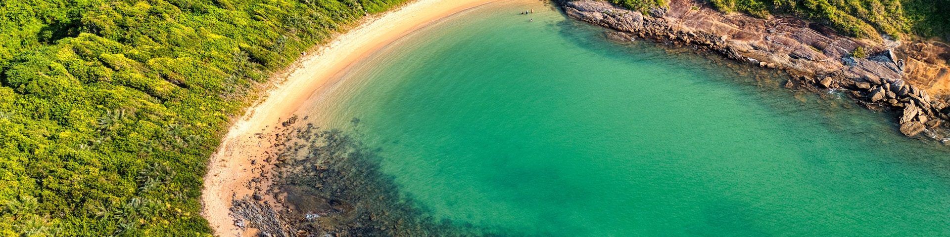 Vista aérea de uma área costeira com praia de areia, águas turquesa cristalinas e vegetação verde exuberante.