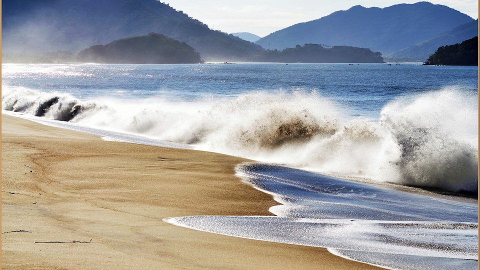 A beach with waves crashing onto the shore, surrounded by mountains.