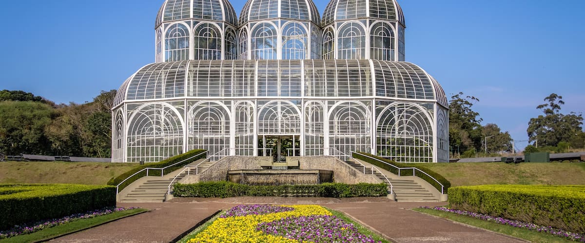 A large greenhouse with multiple domes and arched windows, surrounded by a well-maintained garden with colorful flower beds.