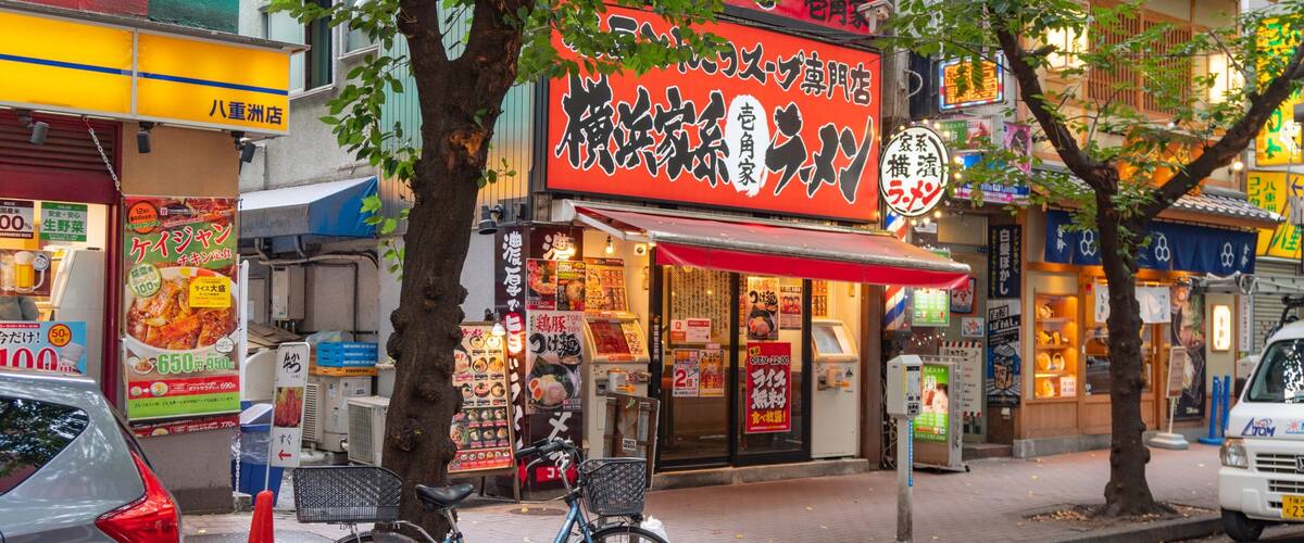 A street with a bicycle parked by a tree, a car, and a ramen shop with a red awning.