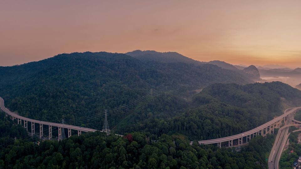 A long bridge curving through a mountainous landscape with dense forests.