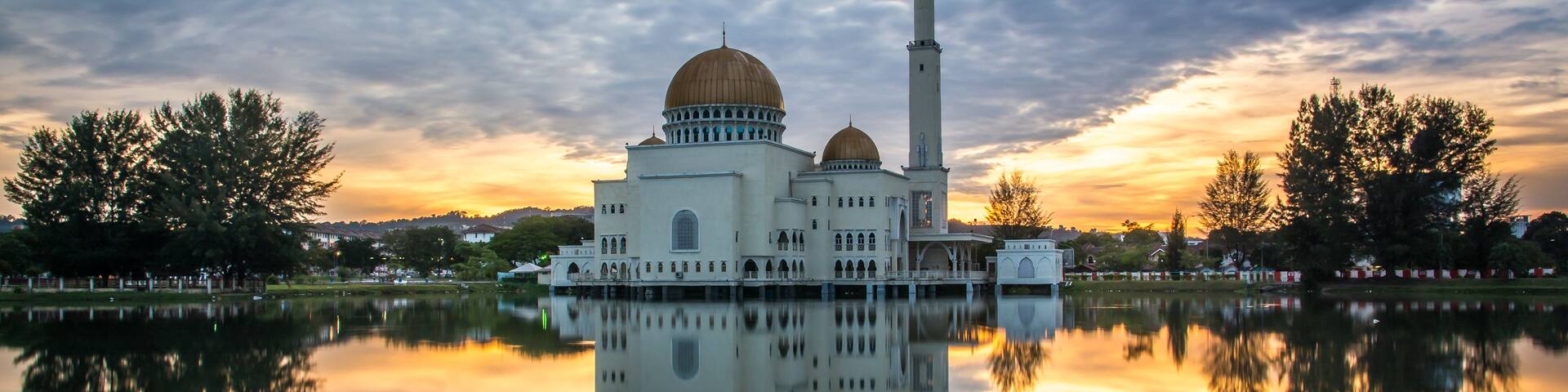 A mosque with a large dome and a tall minaret, reflecting in a calm body of water, surrounded by trees and a sunset sky.