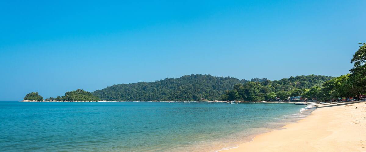 A beach with clear turquoise water, sandy shore, and a forested hill in the background.