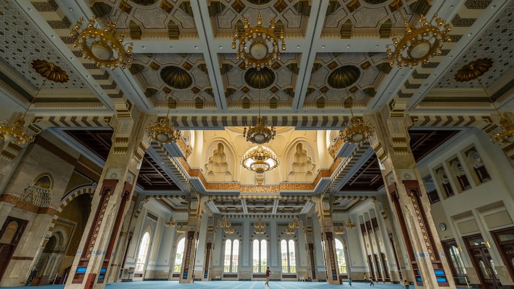 Ein kunstvoll gestaltetes Moscheeforum mit blauen Gebetsmatten, goldenen Kronleuchtern und aufwendigen geometrischen Mustern an der Decke.