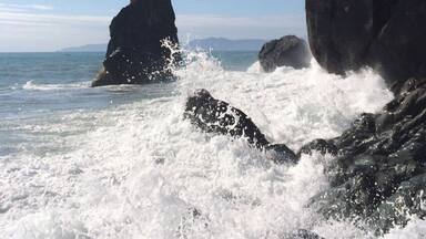 A rocky coastline with waves crashing against large boulders and a prominent rock formation in the distance.