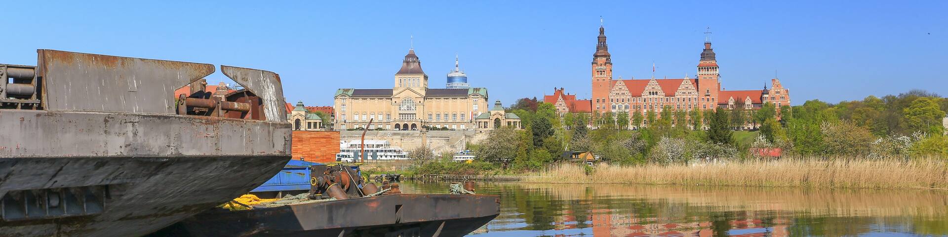 A river with a large ship docked on the left, a historic building with towers in the background, and a clear blue sky.