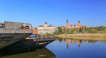 Une rivière avec un grand navire amarré sur la rive gauche, un bâtiment historique avec des tours en arrière-plan, et un ciel bleu dégagé.
