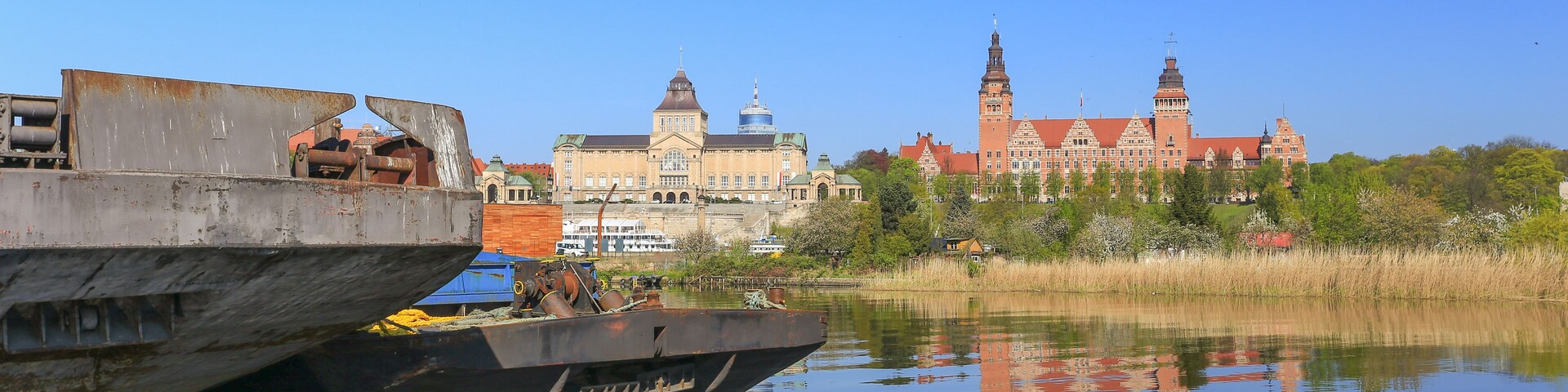 Une rivière avec un grand navire amarré sur la rive gauche, un bâtiment historique avec des tours en arrière-plan, et un ciel bleu dégagé.