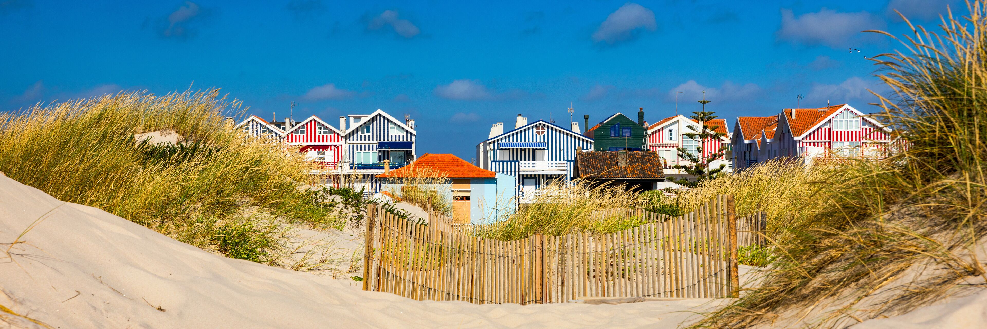 A row of colorful beach houses with sandy dunes and tall grass in the foreground.